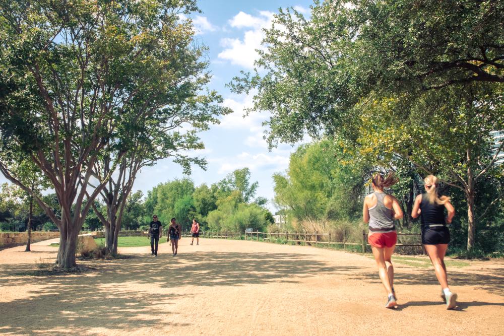 People running and walking along a wide, oak tree-shaded path of the Hike-and-Bike Trail