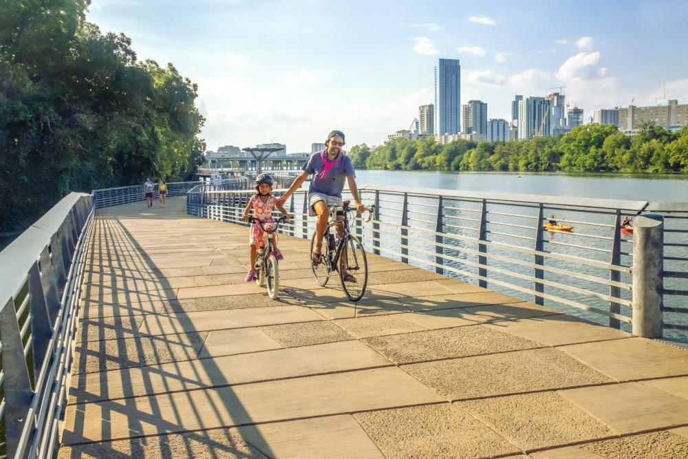 Image of a man and daughter riding bikes on the Butler Hike and Bike trail boardwalk with Austin Downtown in the background.