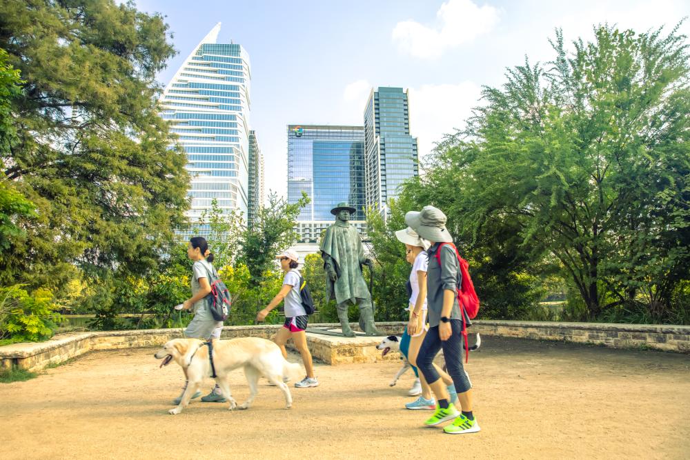 Men and women walking past the Stevie Ray Vaughan statue on the Butler Hike and Bike Trail with their dogs.