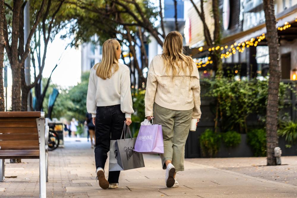 photo shows two women from the back, walking down a sidewalk and holding shopping bags