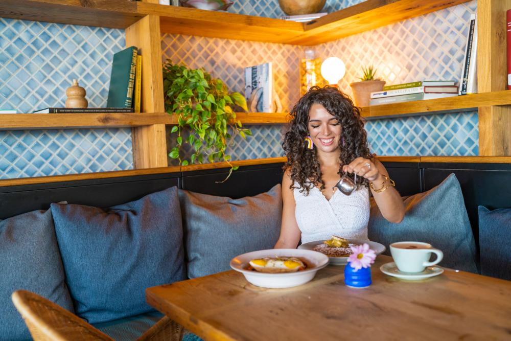 Photo of woman sitting at corner table surrounded by blue cushions in Cafe No Se pouring syrup on pancakes. There is a cup of coffee, a flower and another dish on the table