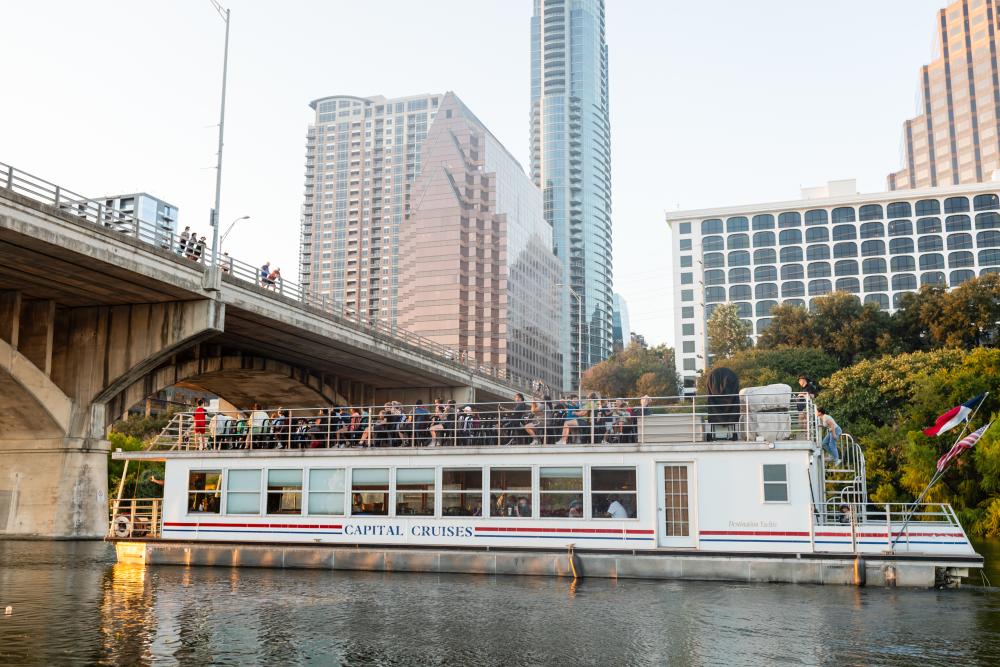 A Capital Cruises boat floating under the Congress Avenue bridge with people standing on the top deck of the boat waiting for the bats to fly out.