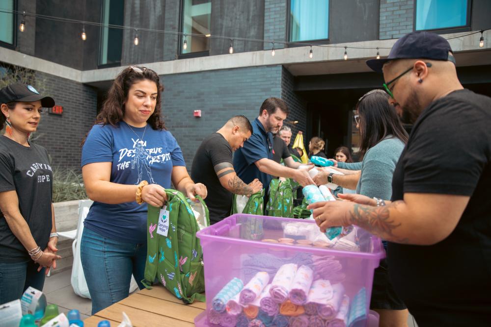 Group of volunteers filling backpacks with care items in an outdoor courtyard.