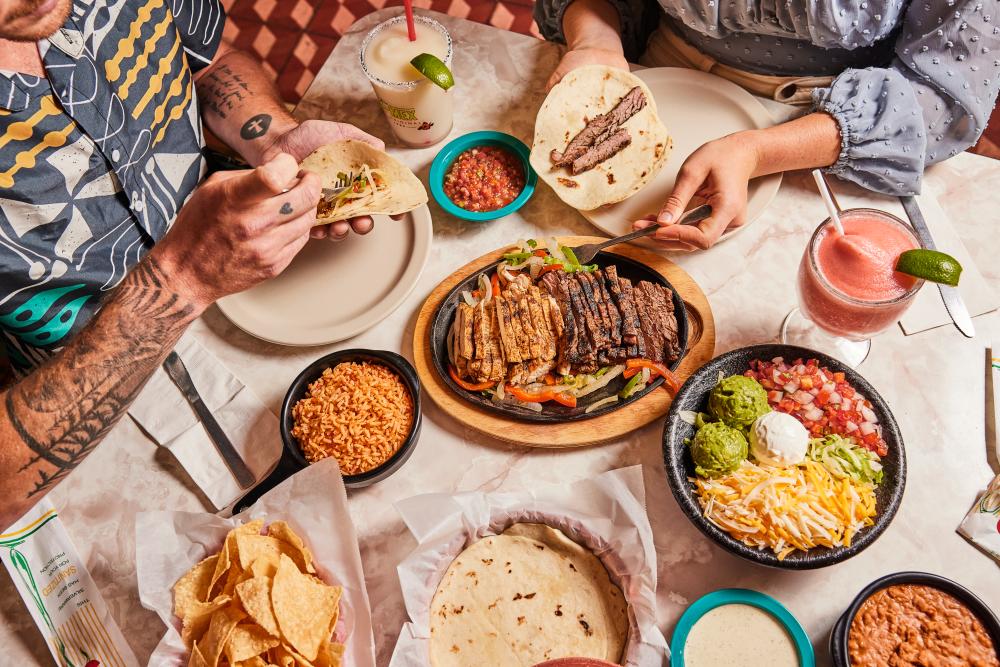 People putting fajita meat on tortillas surrounded by bowls and plates of Tex-Mex.