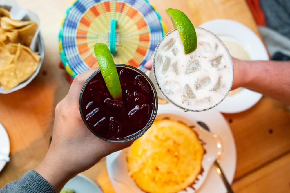 Two hands holding a cocktail and margarita over colorful plates of Tex-Mex.