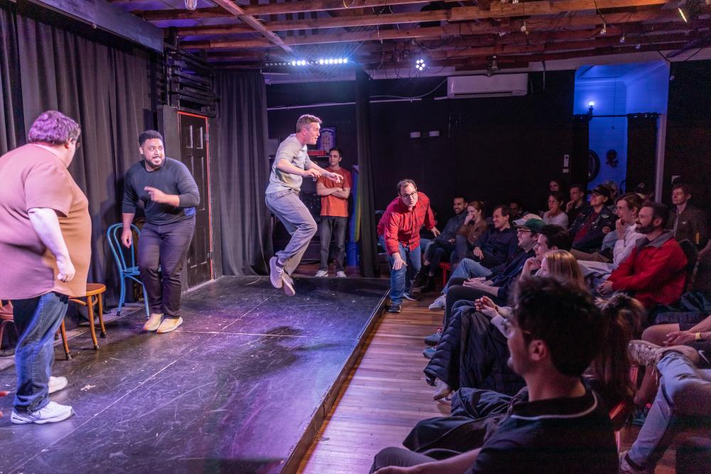 A group of men on stage at ColdTowne Theater doing a performance in front of an audience where one man is mid-jump on stage.