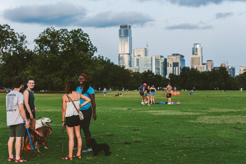 Different groups of people, some with dogs, scattered around green Zilker Park, with downtown skyline in the background.