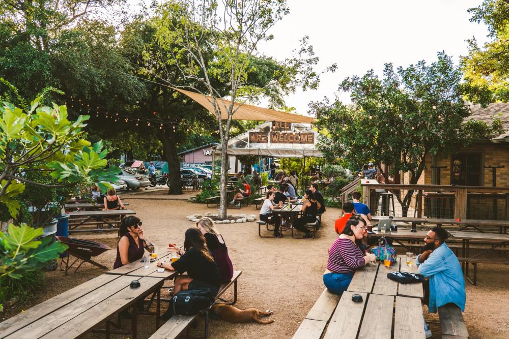 Groups of people sitting at picnic tables in a courtyard surrounded by greenery and a covered patio in the background with the words "ALL ARE WELCOME" posted to the front of the structure.