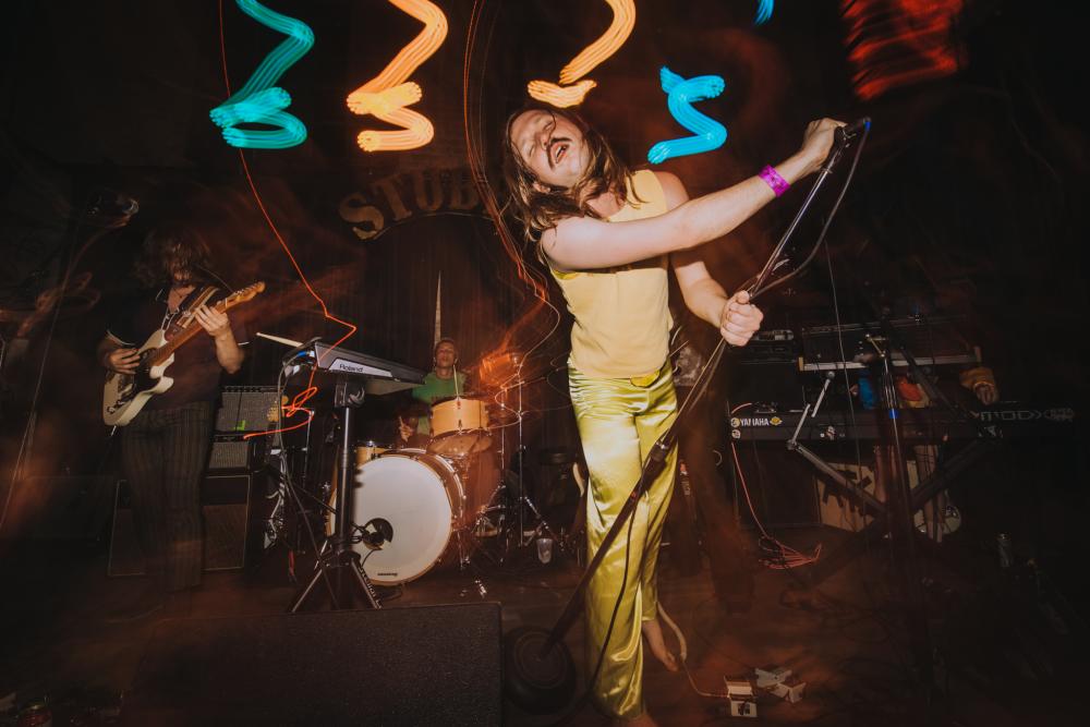 Barefoot man on stage holding a microphone stand, with blurry lighting in the background.