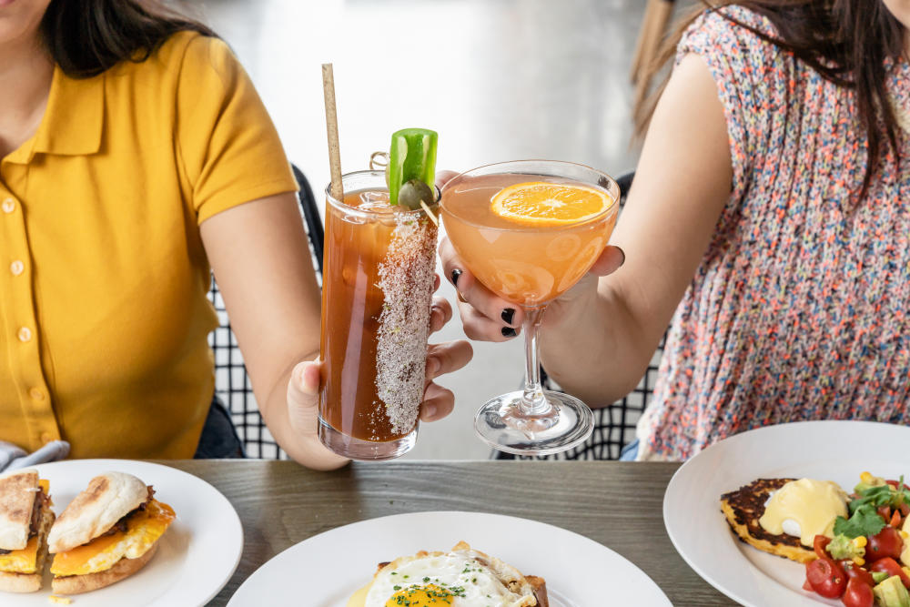 Two women clinking cocktails together over a large breakfast spread.