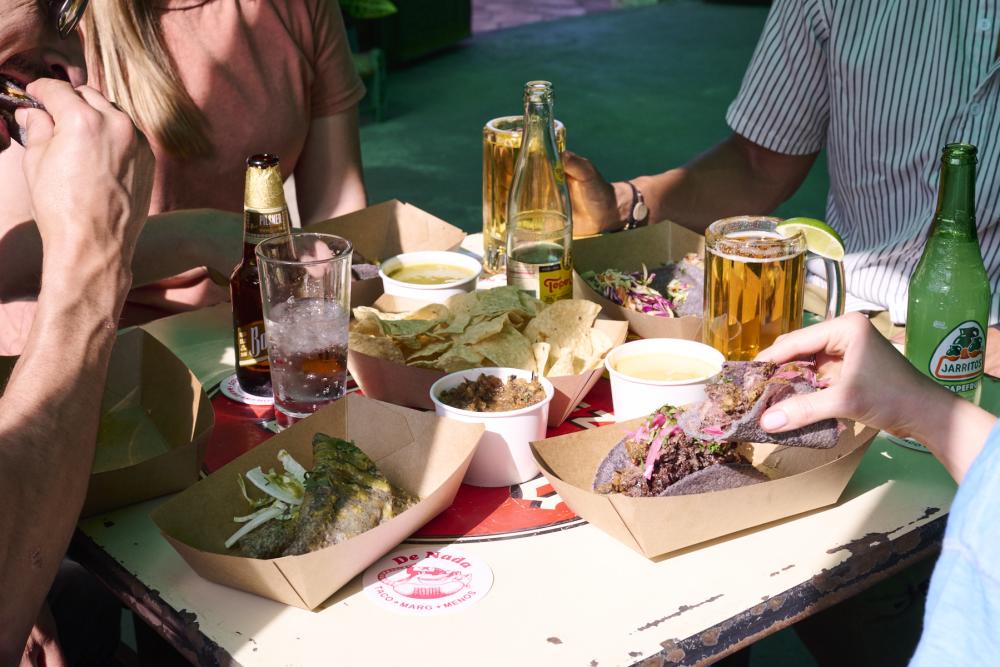 People gathered around a table with cardboard serving boats of tacos, queso and chips.