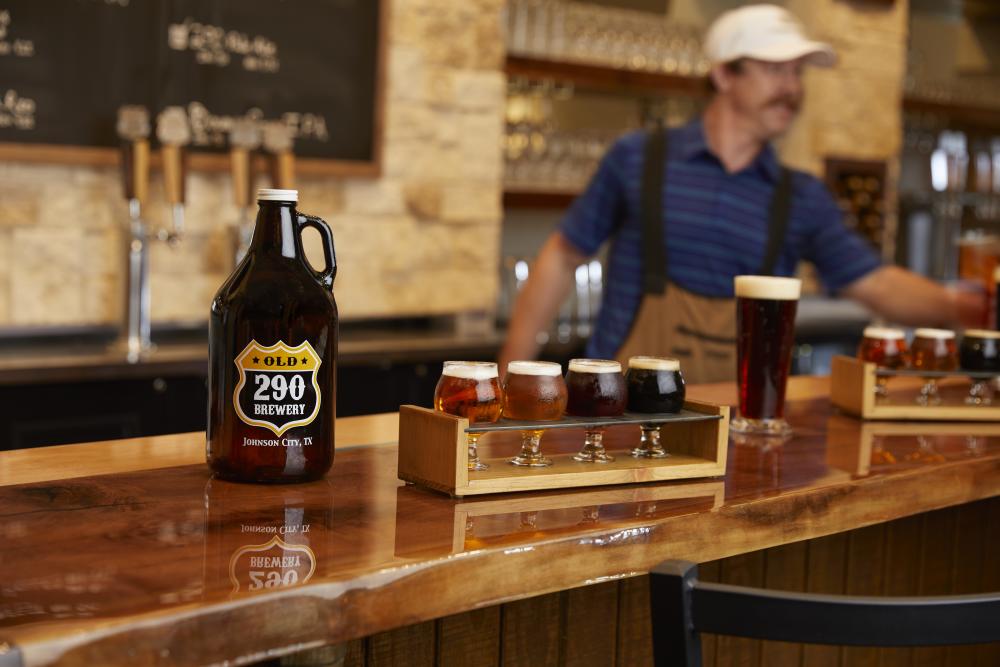 Photo of a beer growler reading Old 290 Brewery Johnson City, TX next to a tasting flight of beer on wooden bar and a bartender in white baseball cap behind the bar at Carter Creek Winery Resort