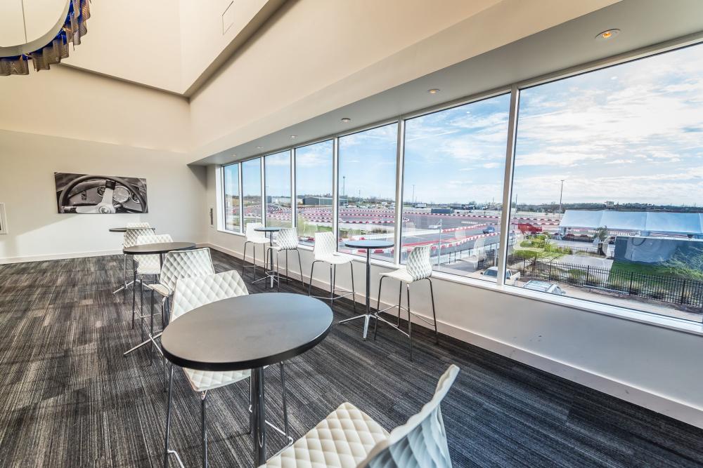 Empty Meeting Room at Circuit of the Americas with five two-top tables in front of a wall of windows. Outside the window, the racetrack is visible