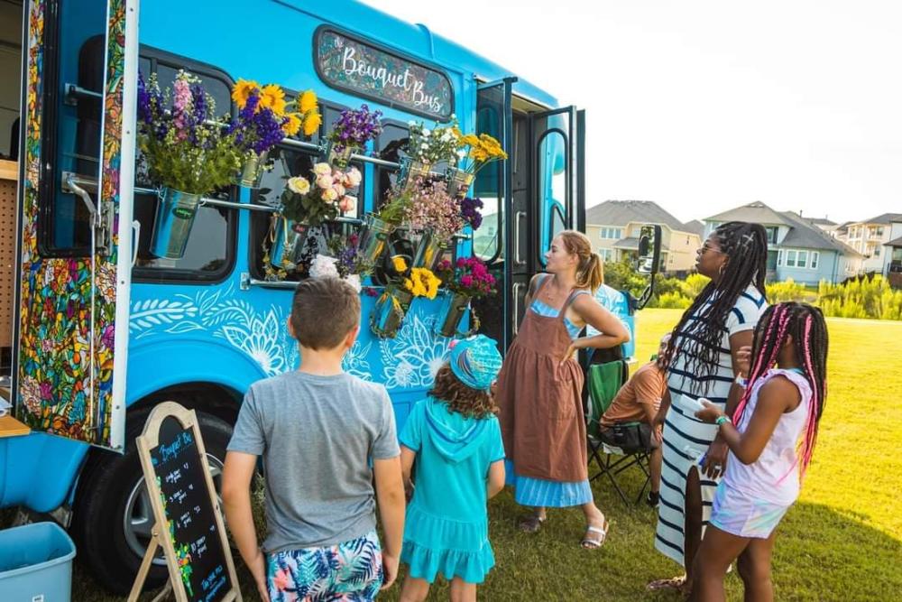 People standing in field outside of a blue bus, with buckets of flowers on the outside.