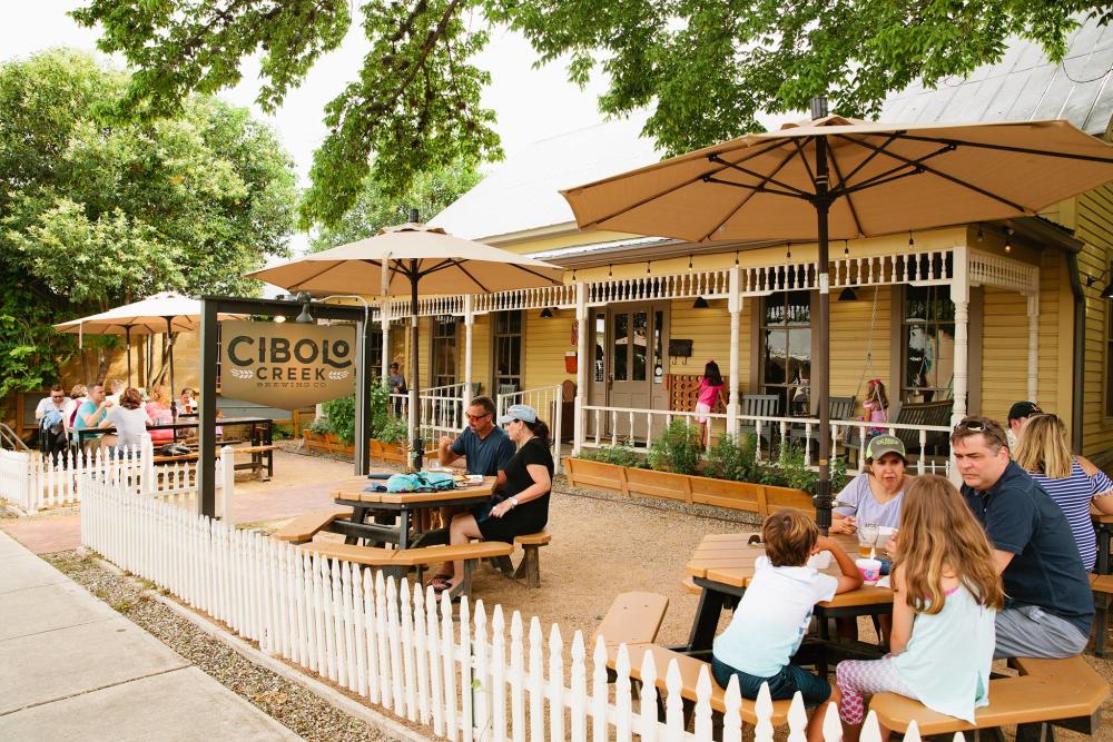 Photo of a busy patio in front of the Cibolo Creek Brewing Co building. Families sit at each of the tables under umbrellas
