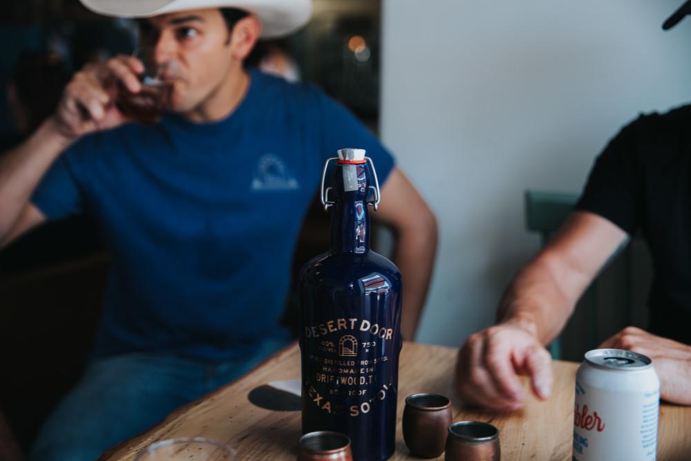 Man in cowboy hat sipping on cocktail with bottle of Sotol on a table.