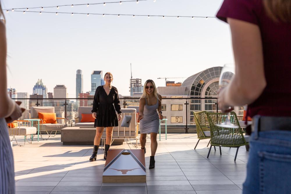 Four women playing cornhole on a rooftop patio overlooking the Austin skyline.