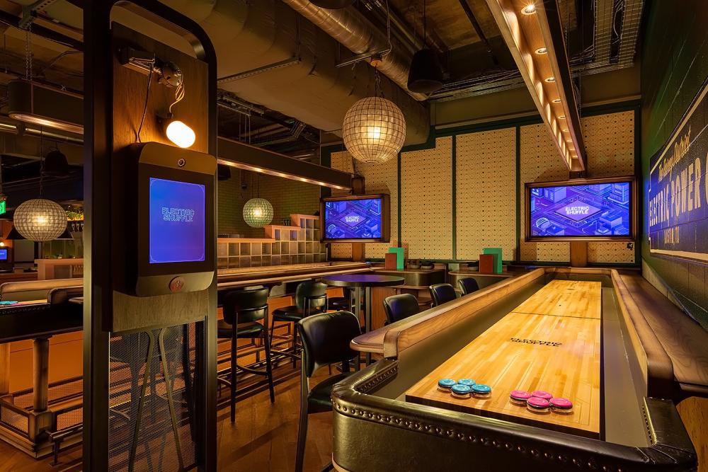 Shuffleboard game surrounded by bar seats, retro decor and TV screens.