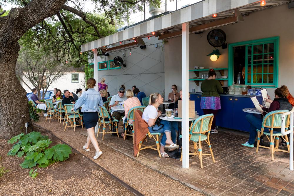 People sitting at bistro tables under a covered patio shaded by a large oak tree.