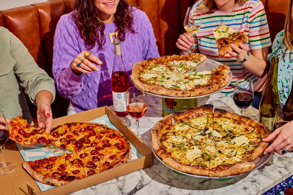 Women sitting in a booth with a table covered by pizza and wine bottles.