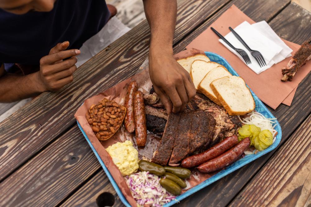 Plastic tray covered in butcher paper topped with slices of different barbecue meats and traditional sides.