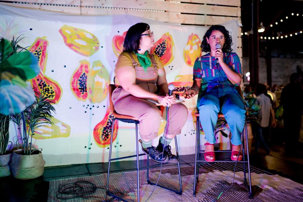 Two women sit on stools and present in front of a colorful mural depicting fruit at the Front Festival & Market