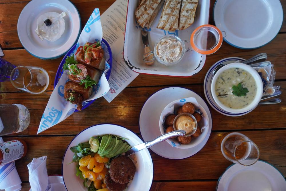 Overhead view of a wooden table covered in plates of lobster rolles, smoked fish dip, hush puppies and more.