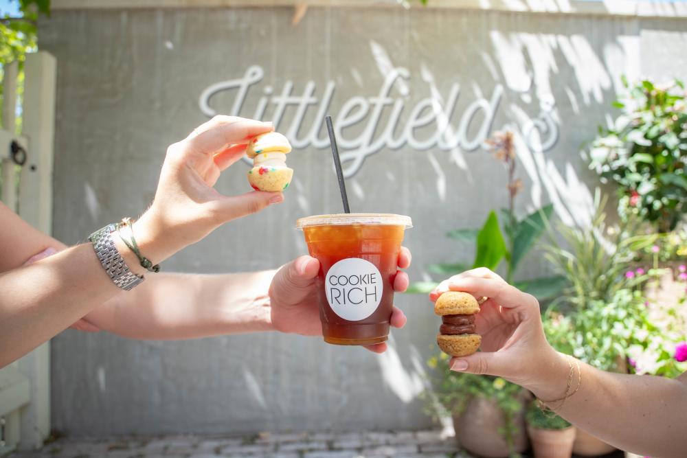 Hands holding up a coffee and little cookies in front of the Littlefield's sign.