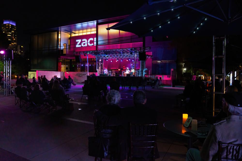 Photo of a performance of Gospel Down By The Riverside ZACH Theatre. The show is being held outside at night, and people are seated at tables in front o the building