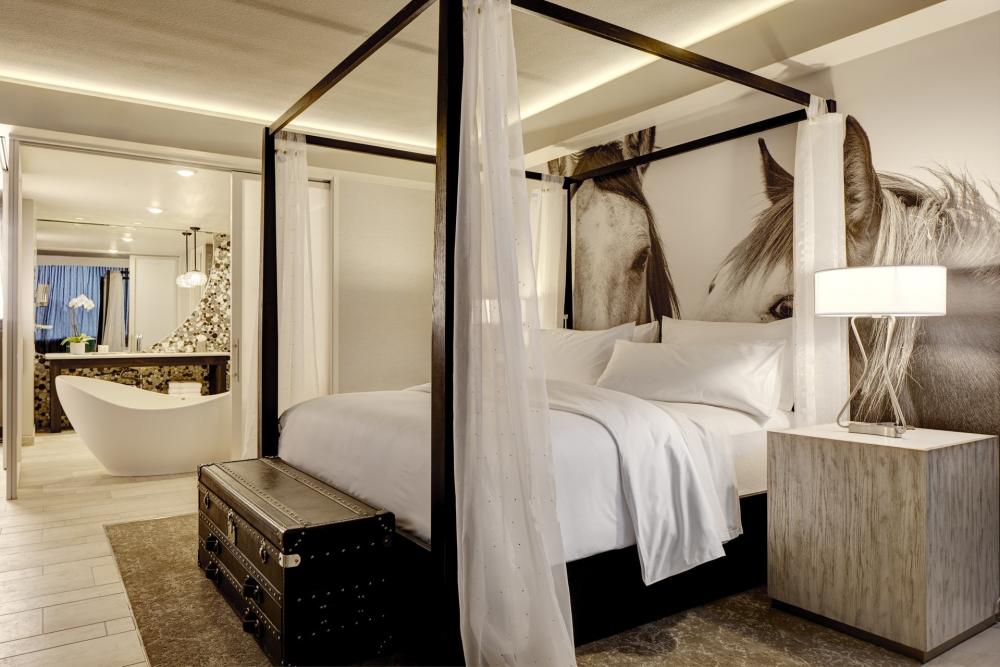 Archer Hotel's King Suite showing a four poster bed with white linens and curtains. A freestanding bathtub is visible in the bathroom in background