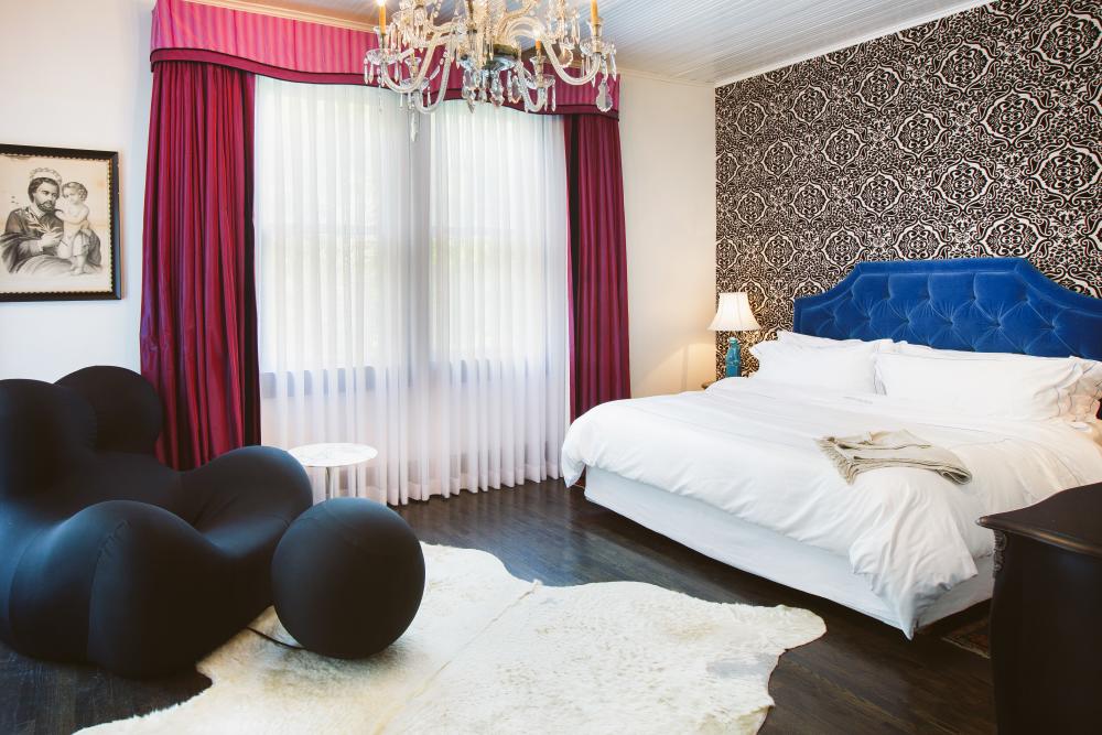 Hotel Saint Cecilia's Suite 4 Bedroom with a large, modern black armchair and pouf, a chandelier, pink and white curtains, a blue headboard and textured wallpaper behind the bed