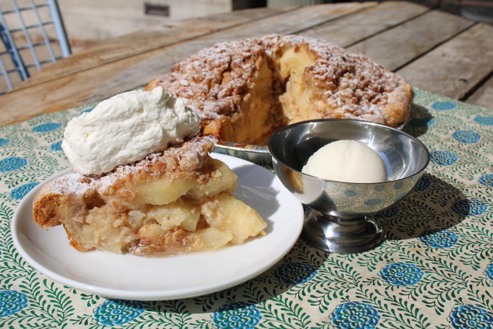 Thick slice of chunky apple pie topped with homemade whipped cream, sitting next to the full pie it was taken from and a silver chalice filled with ice cream.