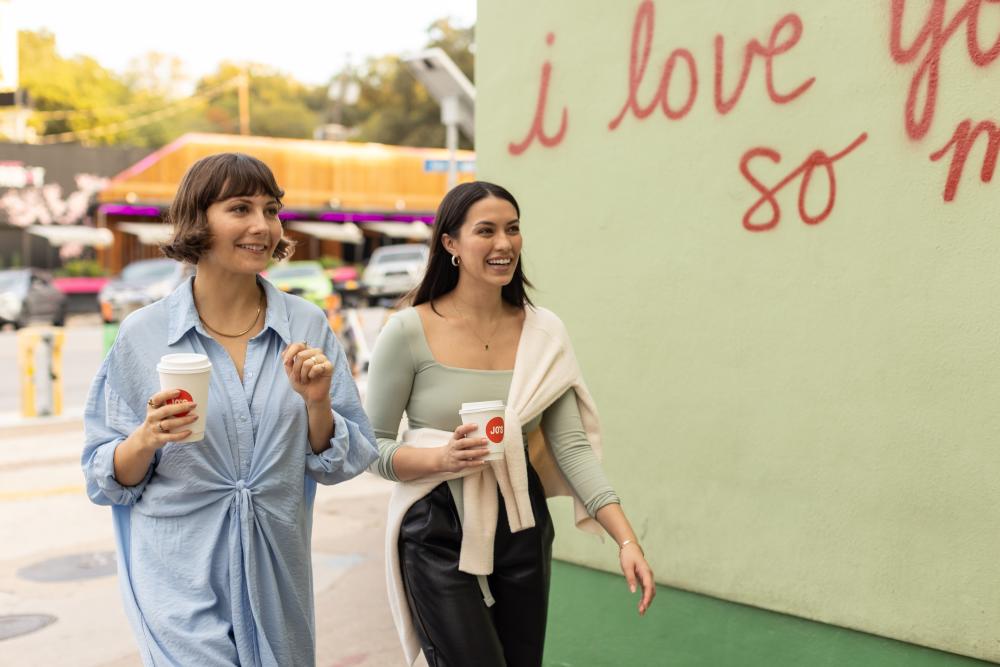 Two women walking past "I love you so much." mural with to-go coffee cups in hand.