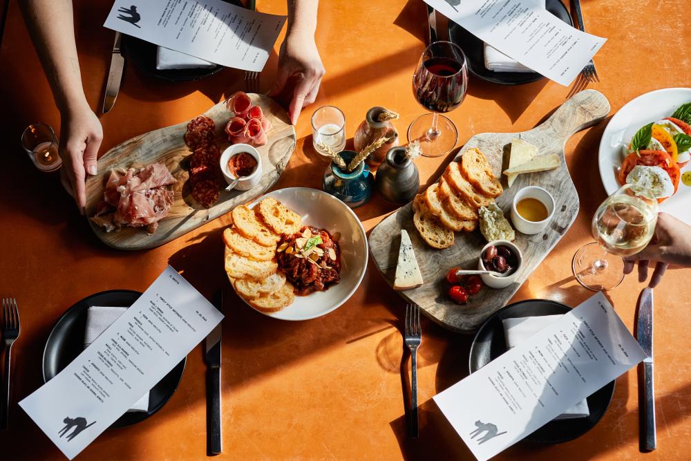 Mixed antipasto plates with breads, dips and accoutrement on an orange table top.