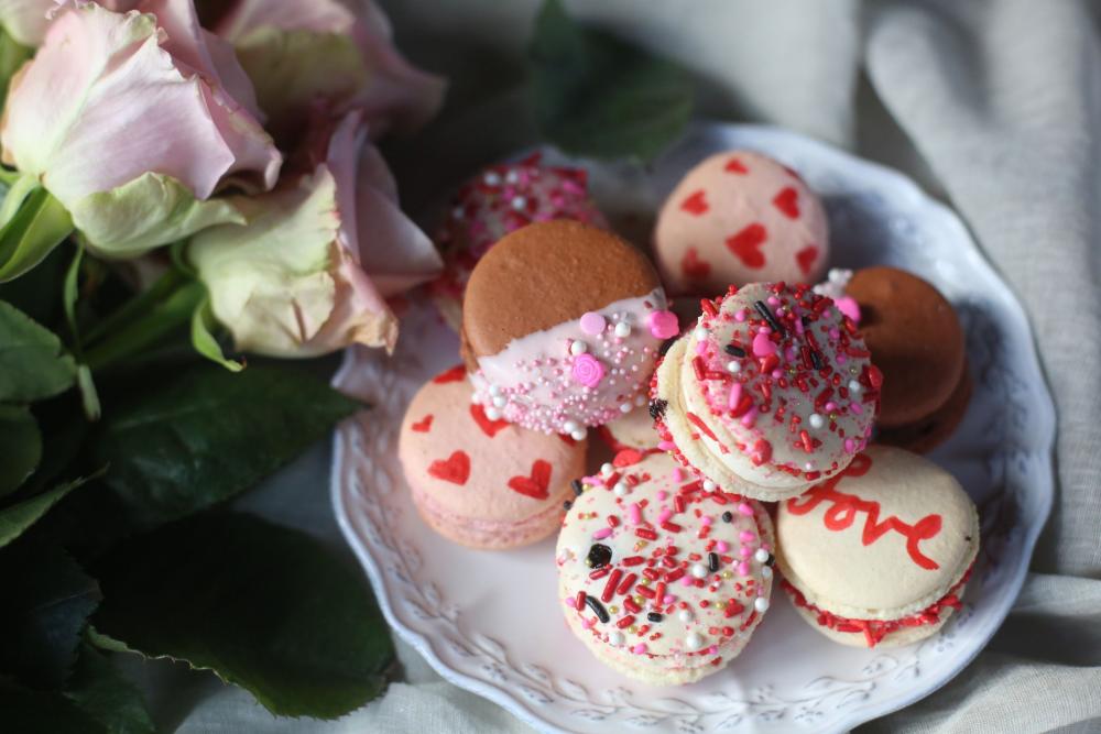 Pink and white Valentine's themed macarons on a decorative plate next to a bunch of roses.