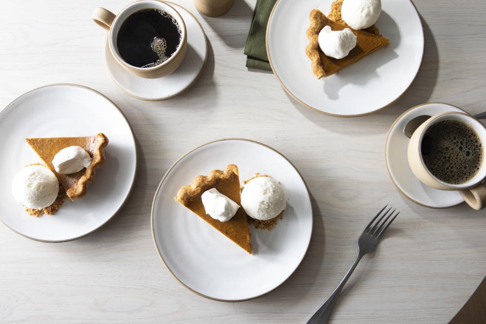 Slices of pumpkin pie and single scoops of vanilla ice cream on white plates, next to steamy cups of coffee.