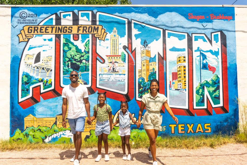 Family of four with two young daughters hold hands and smile at camera in front of postcard mural that reads Greetings from Austin Texas