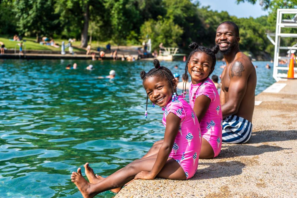 Two little girls and their father sit on the edge of the pool and smile at Barton Springs