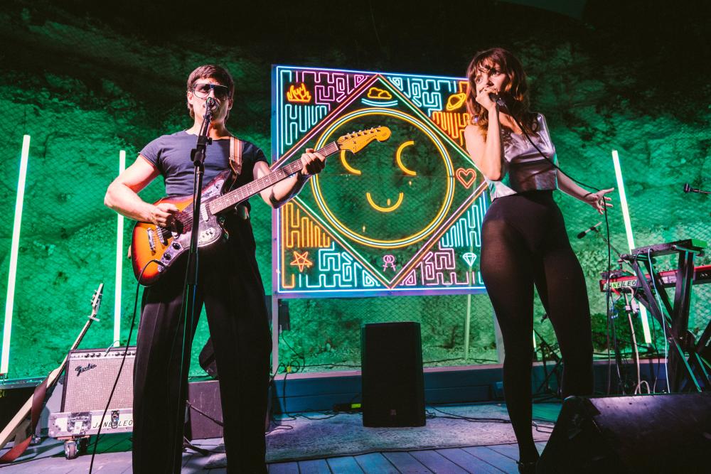 Man playing guitar and woman singing into a microphone on a stage backlit by a neon smiley face.