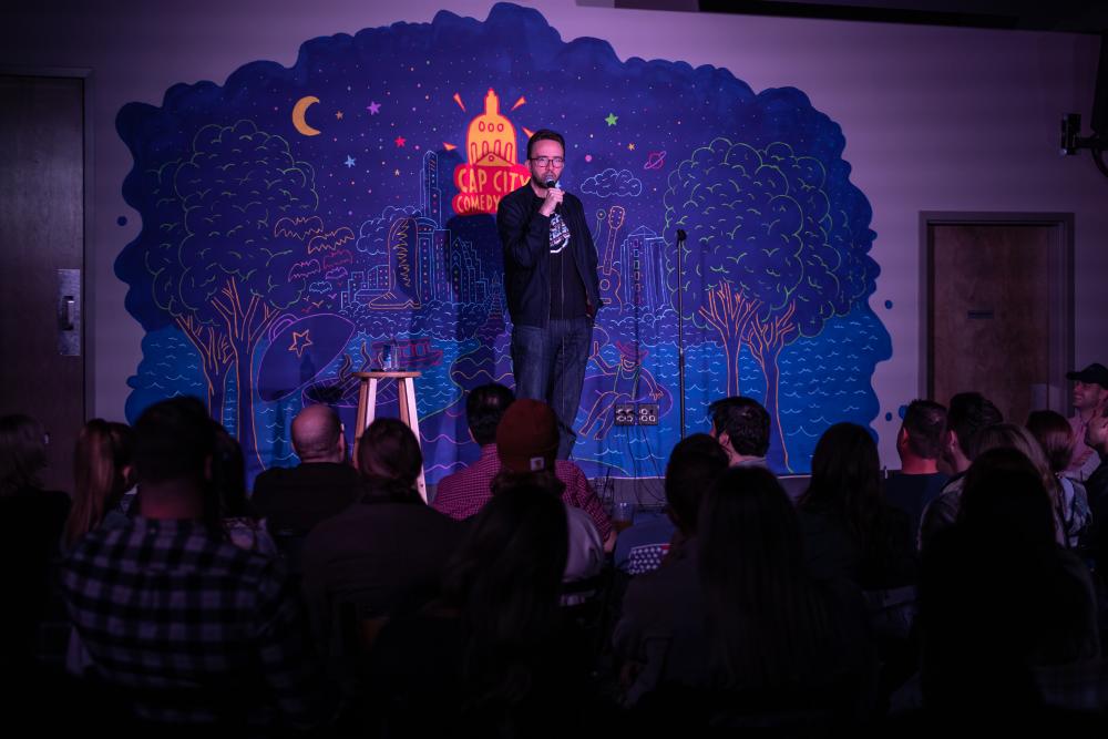 Joe List performing stand-up comedy on the stage of Cap City Comedy Club in front of a shadowy audience.