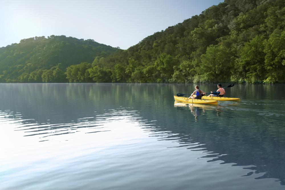 Couple kayaking on glassy water of Lake Austin in between lush, tree covered rolling hills.