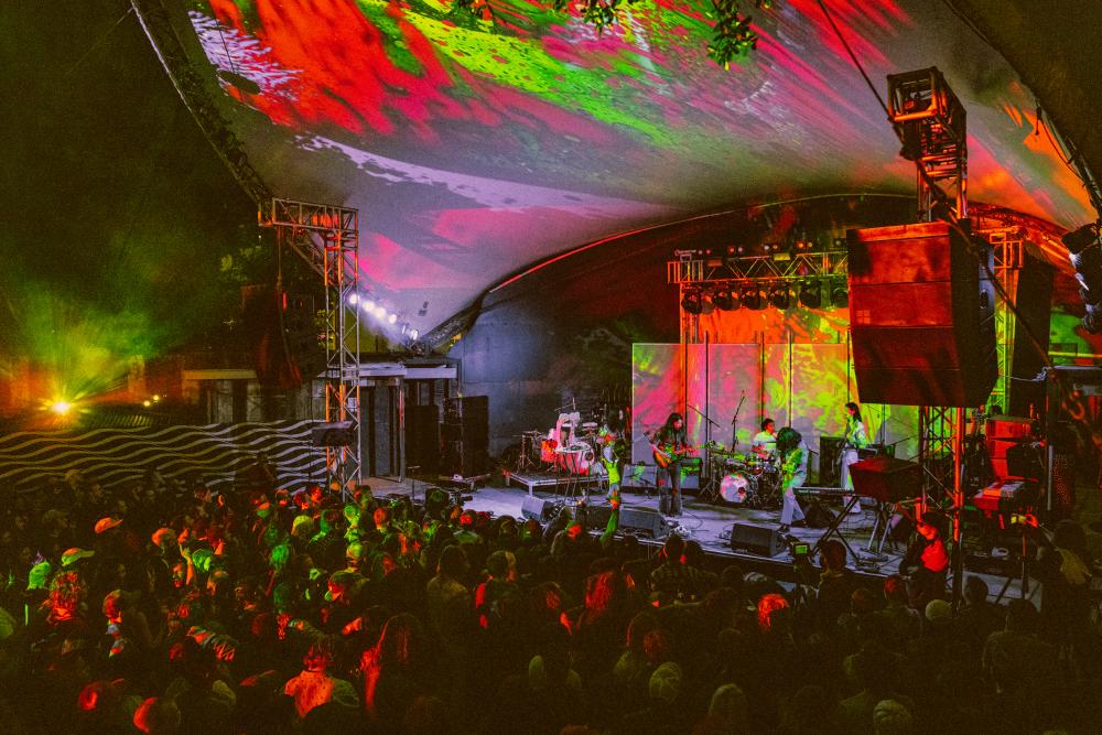 Large crowd standing in front of colorfully lit outdoor stage.