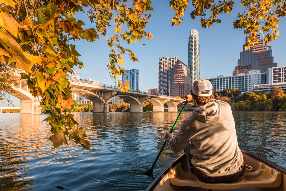 Man in Canoe on Lady Bird Lake across from downtown Austin Texas skyline with fall leaves