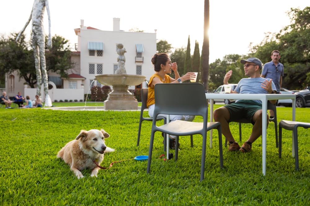 Man and woman sitting with dog in the grassy area of Laguna Gloria, near sculptures.