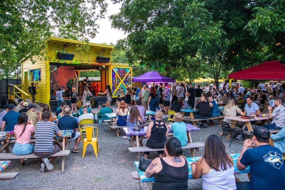 People sitting at outdoor picnic tables listening to a band on an outdoor stage.