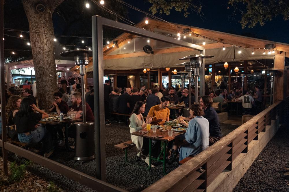 People sitting at picnic tables on a large restaurant patio with string lights and heaters.