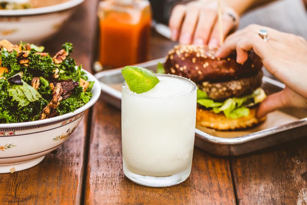 Frozen margarita in a frosty glass next to a bowl of kale salad and a sizeable burger, on a patio table.