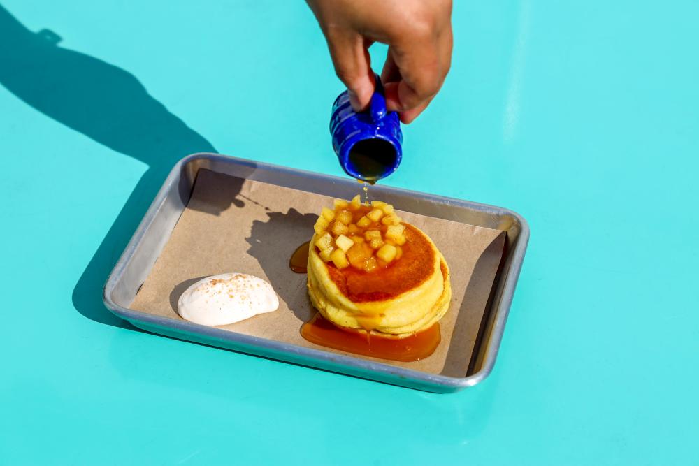Stack of mini, fluffy pancakes sitting on a metal eighth sheet pan, topped with diced apples, drizzled with syrup.