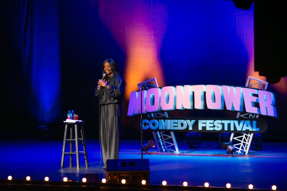 Image of a woman standing on-stage with a mic in her hand with a sign reading "Moontower Comedy Festival" sitting behind her.