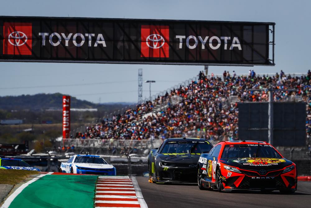 Cars winding down a racetrack with stands full of fans in the background.